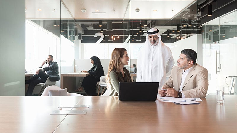 A photo of multi-ethnic Arab, Caucasian and Middle Eastern business people discussing at conference table in modern office. Professionals are with laptop. Some are in formals and others are wearing traditional attire. Dubai, United Arab Emirates.